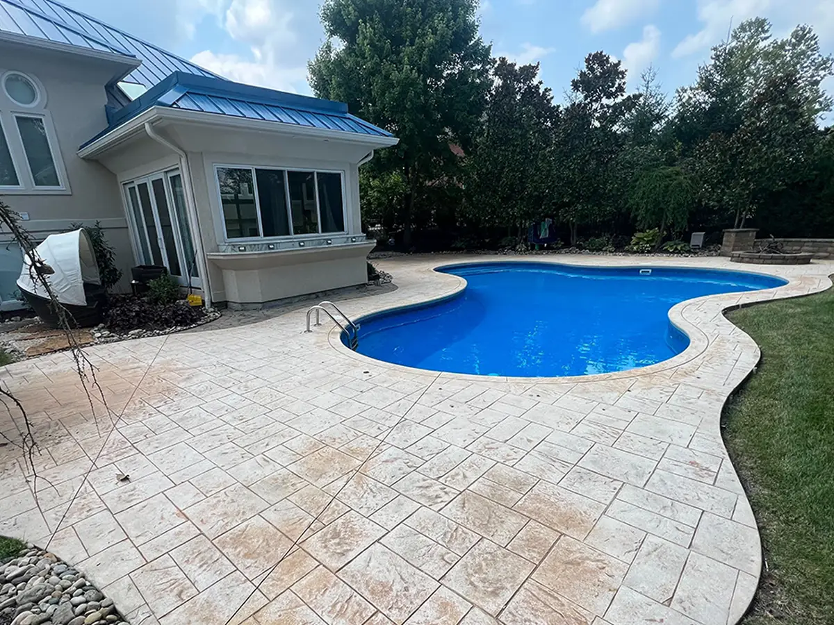 Stamped concrete patio surrounding a blue swimming pool, showcasing the textured surface and landscaping, emphasizing the need for sealing to protect against elements.
