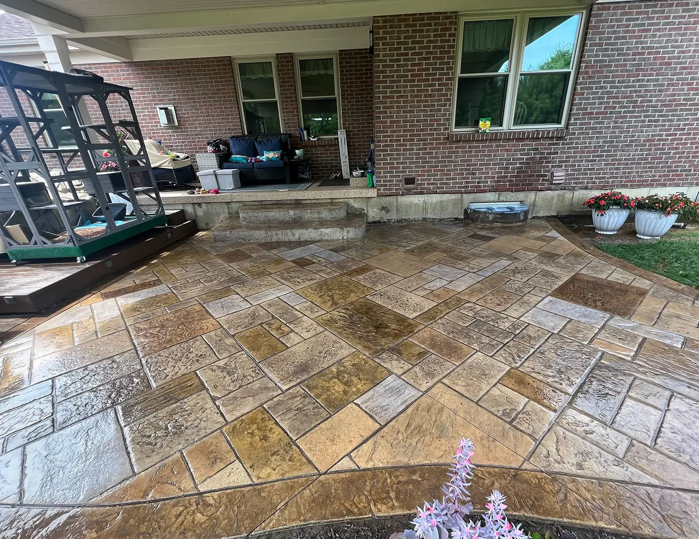Stamped concrete patio with a recently sealed surface, showcasing a blend of rich colors and textures, surrounded by a residential setting, including a porch and flower planters.