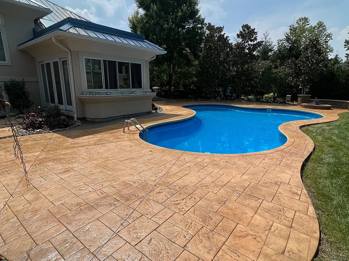 Stamped concrete pool deck surrounding a blue swimming pool, showcasing a textured surface with a seamless transition to a landscaped area, emphasizing the aesthetic appeal and durability of professional sealing services by Perfect Paver Co.