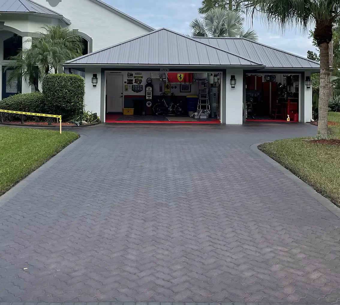 Paver driveway leading to a garage, showcasing a vibrant, restored surface with a herringbone pattern, surrounded by landscaping, illustrating the benefits of professional paver tinting services by Perfect Paver Co.