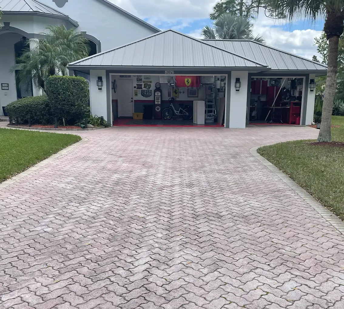 Paver driveway leading to a garage with a red accent, showcasing faded pink pavers in need of restoration, surrounded by greenery and a clear sky, illustrating the potential for vibrant outdoor surface transformation.